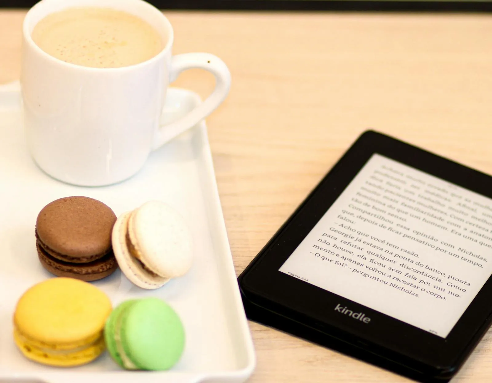 Kindle Paperwhite com texto em português ao lado de uma chávena de café e um pratinho com macarons coloridos