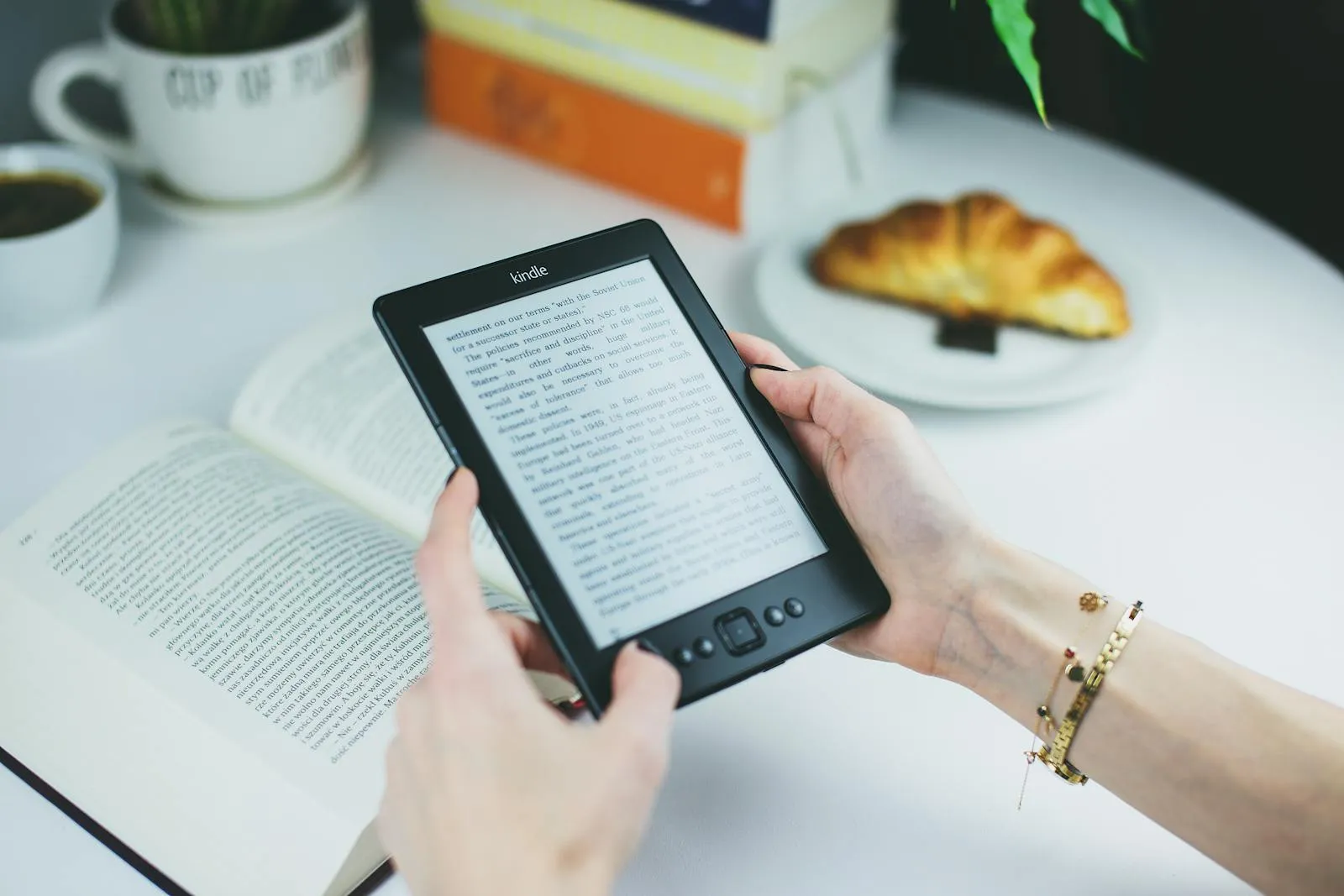 Mãos a segurar um Kindle sobre um livro de papel aberto, com café e croissant ao fundo