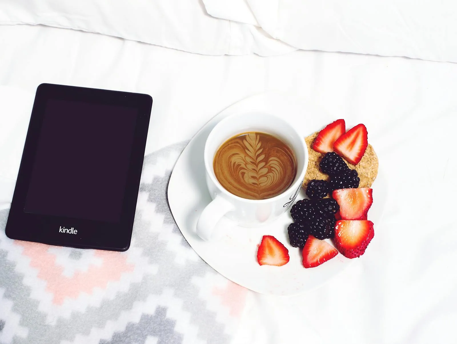 Kindle Paperwhite sobre lençóis brancos, ao lado de um café com latte art e frutos vermelhos